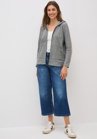 Grau zip-up Hoodie mit Kapuze und Kordelzügen, kombiniert mit weit geschnittenen blauen Denim-Jeans und weißen Sneakern mit schwarzen Akzenten.
