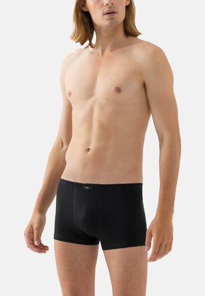 Oberkörperfreier männlicher Model mit heller Haut und schulterlangen Haaren, das schwarze Boxershorts trägt und vor einem schlichten hellen Hintergrund steht.