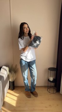 Oggetto nero in un sacchetto di plastica tenuto da una persona con una maglietta bianca e jeans blu, in piedi in una stanza con pareti di colore chiaro e pavimento in legno.