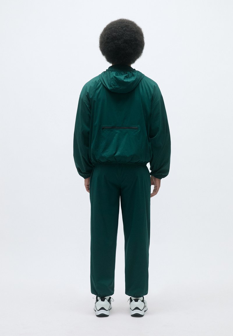 Dunkelgrüne Sportjacke mit Kapuze und elastischen Bündchen, kombiniert mit passenden Hosen. Das Outfit verfügt über eine mit Reißverschluss verschlossene Tasche auf der Rückseite.