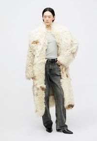 Παλτό από shearling σε κρεμ απόχρωση με καφέ λεπτομέρειες, oversized σχεδίαση, υφή με ανάγλυφη επιφάνεια, φορεμένο πάνω από γκρι μπλούζα και συνδυασμένο με γκρι τζιν παντελόνια με φαρδιά μπατζάκια.