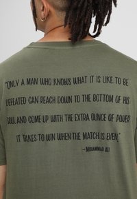 Camiseta de algodón en color verde oliva con texto negro en la parte de atrás que incluye una cita atribuida a Muhammad Ali. Diseño simple y sin adornos.