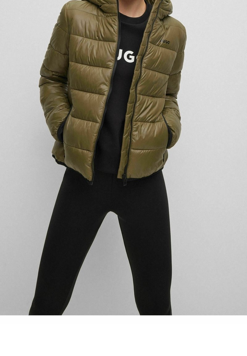 Giacca puffer verde oliva con cappuccio, chiusura con zip e trapuntatura orizzontale. Presenta un logo sul petto sinistro, abbinata a una maglietta nera e leggings.