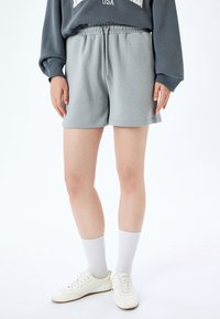 Pantaloni corti in pile grigio chiaro con cintura elastica e cordino. Abbinati a sneaker bianche e calze al ginocchio, su uno sfondo semplice.