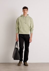 Sudadera con capucha de color verde claro con cordones ajustables, combinada con jeans negros y zapatillas verde oliva. El modelo sostiene una bolsa de tote negra.