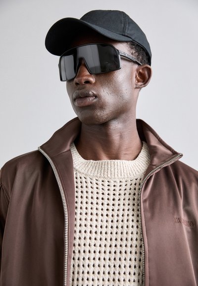 Brun zip-up jakke over en strikket kremfarget genser, kombinert med svarte oversized solbriller og en svart baseballcaps. Glatt stoff og strukturert form.