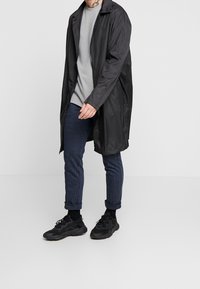 Svart vattentålig rock med krage, lång längd och dragkedja framtill, bärs över en grå texturerad tröja och blå mönstrade byxor. Svarta sneakers.