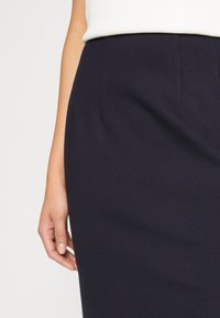 Námořnická sukně-pencil z látky s hladkou texturou, se slim fit střihem a pasem. Viditelná ruka držící okraj sukně.
