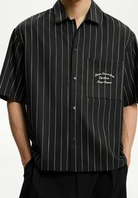 Černá krátká rukávová košile s tenkými bílými vertikálními pruhy, zapínání na knoflíky vpředu a kapsou s vyšitým textem. Hladká, lehká tkanina.