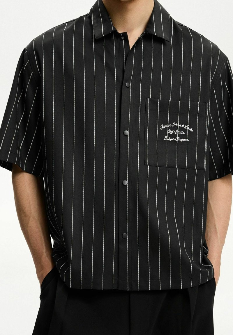 Černá krátká rukávová košile s tenkými bílými vertikálními pruhy, zapínání na knoflíky vpředu a kapsou s vyšitým textem. Hladká, lehká tkanina.