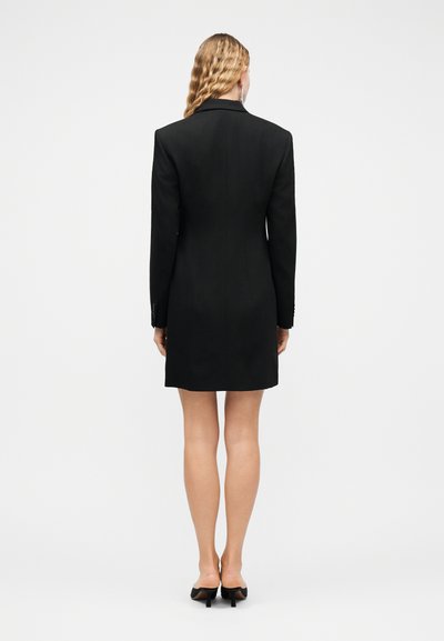Γυναίκα με κυματιστά ξανθά μαλλιά που φοράει εφαρμοστό μαύρο σακάκι-φόρεμα και μαύρα παπούτσια με τακούνια, στέκεται γυρισμένη ανάποδα σε απλό λευκό φόντο.