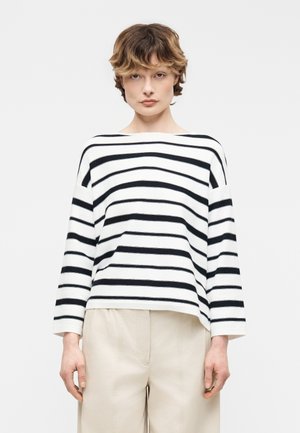 Fiatal személy rövid barna hajjal, hosszú ujjú fehér inget visel fekete vízszintes csíkokkal és bézs nadrágot.