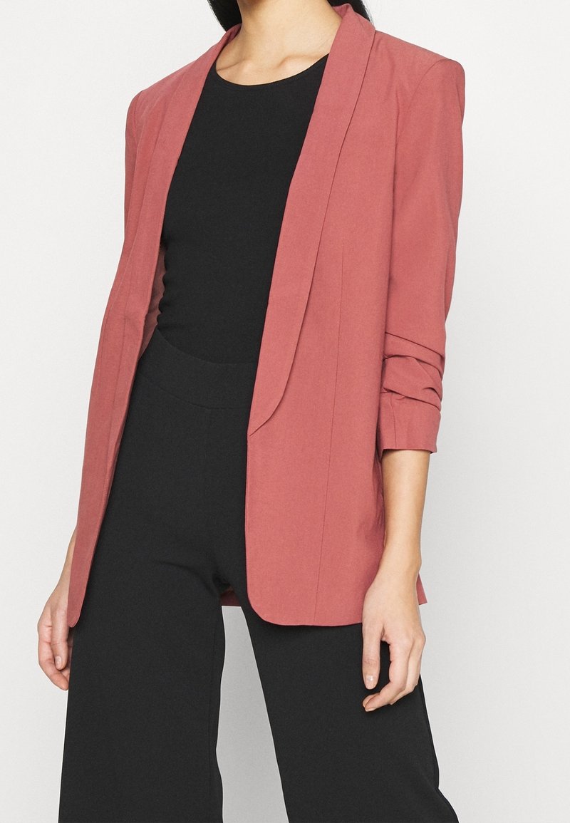 Blazer rosa con un design sartorial, texture liscia e maniche arrotolate, indossato sopra una maglietta nera e abbinato a pantaloni neri a gamba larga.