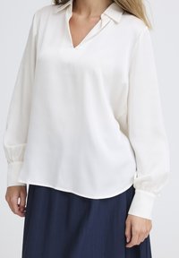 Blusa a maniche lunghe color crema con scollatura a V e vestibilità ampia. Presenta un colletto classico e polsini arr gathered. Tessuto liscio; si abbina a una gonna blu navy.