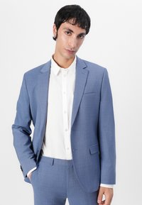 Lichtblauwe blazer van textuurstof, met een enkele knoopsluiting, paspelrevers en een borstzak. Gegdraag over een wit overhemd met knopen.