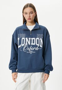 Sudadera de color azul marino con cuello de cremallera de un cuarto, que presenta texto y gráficos en blanco: "LONDRES 1998 2002 Oxford Street West End de Londres Reino Unido."