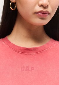Κοντινό πλάνο μιας γυναίκας που φοράει κοραλλί μπλούζα GAP και χρυσό κρίκο σκουλαρίκι, που δείχνει τα χείλη της, το πιγούνι, το λαιμό και μέρος από τα σκούρα μαλλιά της.