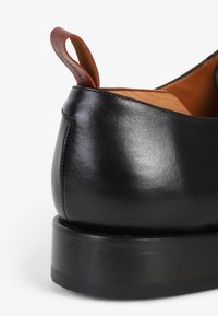 Korkokenkä mustasta nahasta, jossa on sileä pinta, minimaalinen muotoilu ja kontrastiväri ruskea nahkavetoketju. Tasapohja kumista.