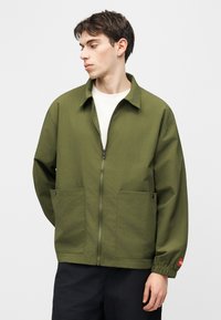 Νεαρός άνδρας που κοιτάζει προς το πλάι φορώντας μια ζακέτα σε ελιά πράσινο με φερμουάρ και μεγάλες μπροστινές τσέπες, καθώς και μαύρο παντελόνι, μπροστά σε απλό φόντο.