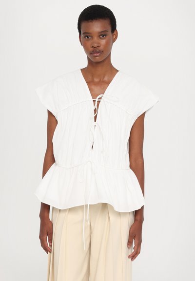 White top with a gathered bodice, wide cap sleeves, and a drawstring tie at the neckline. Textured fabric and peplum hem detail.