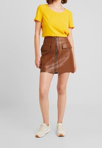 Mini-jupe en cuir brun avec une fermeture éclair à l'avant, deux grandes poches avant, associée à un haut à manches courtes jaune et des baskets beiges.