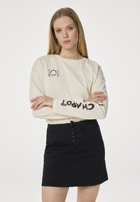 Langarmige crème top met groen en zwarte tekstdetails; gecombineerd met een zwarte rok met een vetersluiting aan de voorkant en een stevige textuur.
