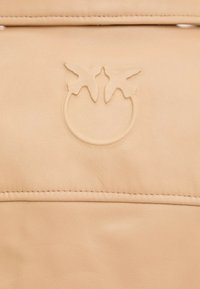 Béžový kožený povrch s vyrazeným logom dvoch vtákov otočených tvárami k sebe nad kruhovým obrysom, viditeľné prešité okraje.