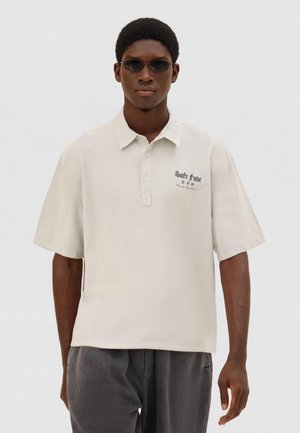 Young man wearing black sunglasses, beige short-sleeve polo with "Soft Fade" text, and dark gray pants against a plain white background.