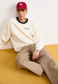 Béžová mikina s čiernym golierom a logom Converse, v kombinácii so svetlohnedými nohavicami. Červená baseballová čiapka s bielym logom NY. Mäkké textúry.