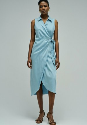 Mujer con vestido cruzado sin mangas de color azul claro con cuello, atado a la cintura, y sandalias marrones de tacón alto, de pie frente a un fondo gris liso.