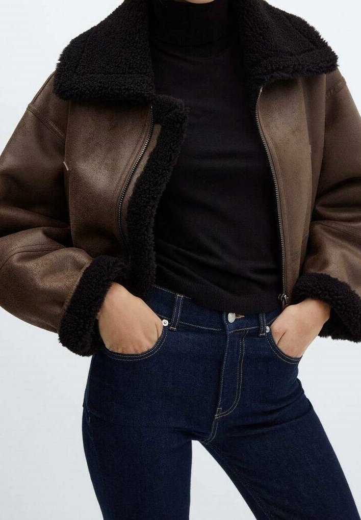 Femme portant une veste en shearling marron avec une doublure noire, un pull à col roulé noir et un jean bleu foncé, mains dans les poches.