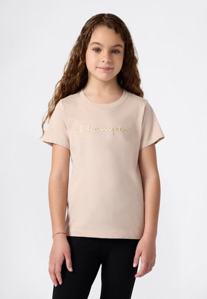 Jeune fille aux longs cheveux bouclés, portant un t-shirt beige Champion et un pantalon noir, se tenant devant un fond clair uni.