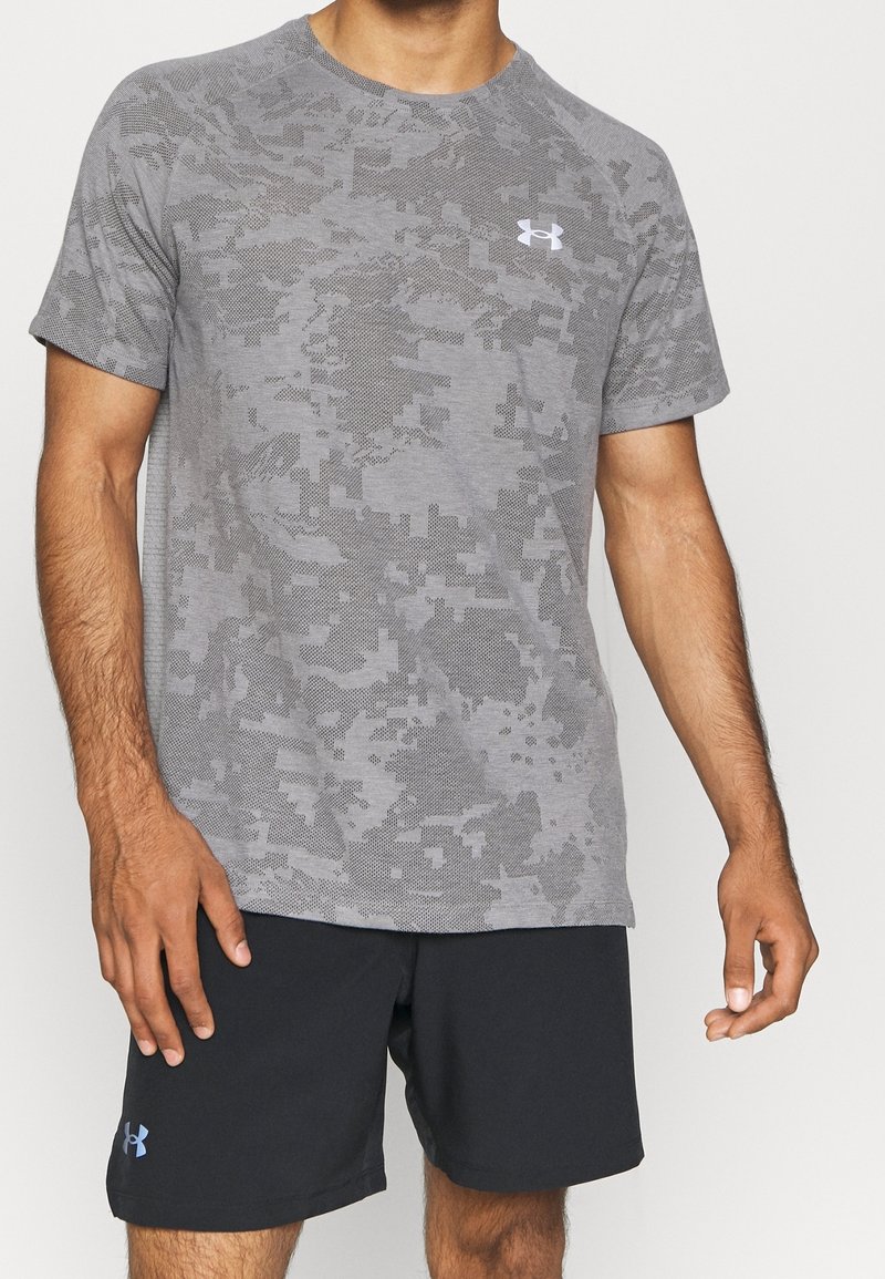 Camiseta atlética gris con patrón de camuflaje, de manga corta, cuello redondo y pequeño logo blanco en el pecho, combinada con pantalones cortos negros.