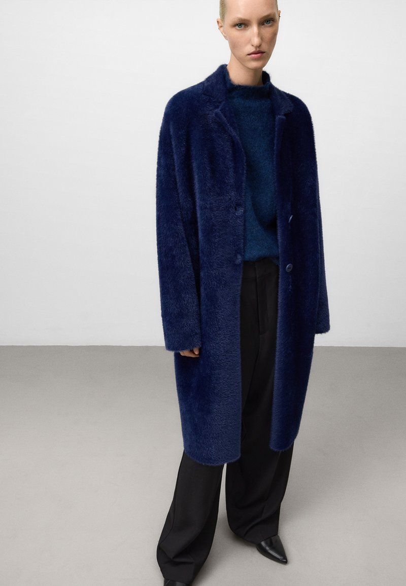 Navyblauer Fake-Fur-Mantel mit Reverskragen, zwei Knöpfen und langen Ärmeln, kombiniert mit einem dunkelblauen Pullover und schwarzen, weit geschnittenen Hosen.