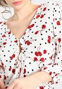 Blouse blanche avec imprimé cerises rouges et pois noirs. Caractérisée par un col en V, un devant froncé avec cordon de serrage et des poignets élastiqués.