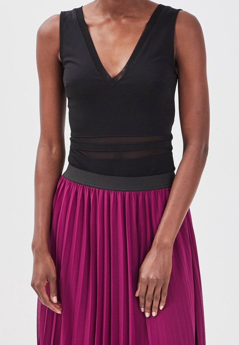 Haut sans manches noir avec un décolleté en V et des panneaux transparents, associé à une jupe plissée magenta et une ceinture noire.
