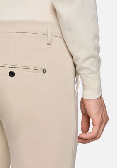 Pantaloni beige su misura con una texture liscia, dotati di una tasca posteriore con zip e un dettaglio con bottone. Mano appoggiata sulla vita.