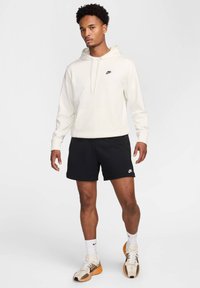 Felpa bianca con cappuccio e tasca frontale, logo Nike nero. Pantaloni corti neri, calze bianche e scarpe da ginnastica con accenti beige e arancione.