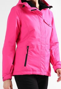 Světlá růžová bunda s kapucí a černými zipovými kapsami, s kapsou na hrudi se klopou, se nastavitelnými zápěstními popruhy, nošena přes černé kalhoty.