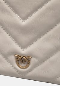 Borsa a tracolla in finta pelle color crema con texture a chevron trapuntata e un logo di un uccello in tono oro sul fronte. Accenti cuciti visibili.