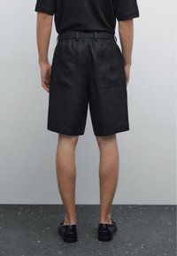 Pantaloni corti neri realizzati in un tessuto liscio, con una cintura elasticizzata e due tasche posteriori, mostrati da dietro su uno sfondo grigio.