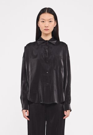 Black, long-sleeve button-up shirt in a smooth, shiny fabric with a collar, front pocket, and relaxed fit. Styled with matching trousers.