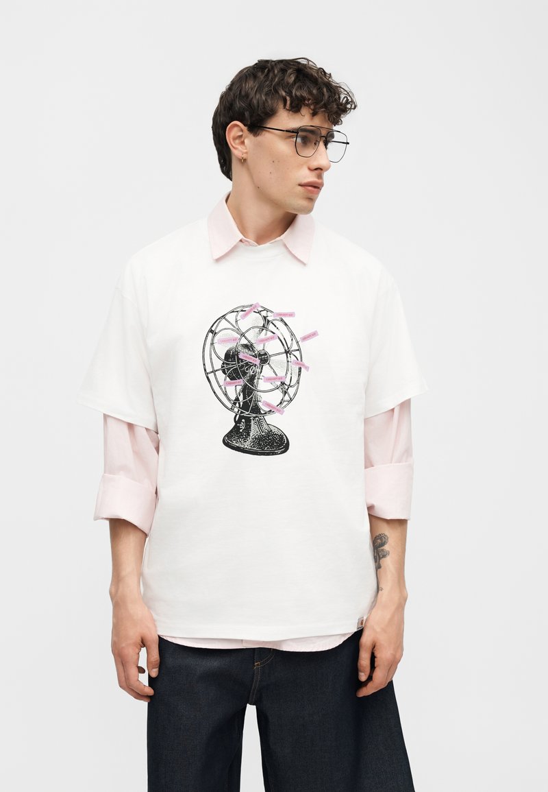 Giovane uomo con capelli ricci e occhiali che indossa una maglietta bianca sopra una camicia rosa, con una grafica nera di un ventilatore vintage con etichette rosa.