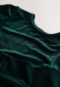 Grønt fløjlstof med en blød tekstur. Har en rund halsudskæring og rynkede detaljer på skuldrene, hvilket skaber et fyldigt udseende.