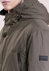 Oliwkowo-zielona pikowana kurtka z wysokim kołnierzem, kieszeniami na zamek i naszywką z logo. Wykonana z teksturowanej tkaniny z trwałymi okuciami.