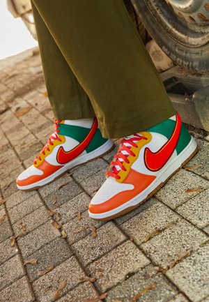 Persona con pantalones verdes y zapatillas coloridas con paneles rojos, naranjas, amarillos, blancos y verdes, de pie sobre una acera pavimentada.