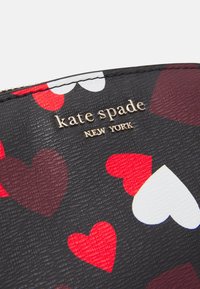 Svart texturerat material med ett mönster av röda, mörkröda och vita hjärtan. Designen inkluderar guldtext med "kate spade NEW YORK"-logotyp.