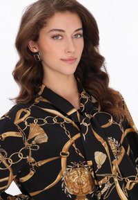 Fekete blúz aranylánccal és díszítő motívumokkal. Kötött gallérral és sima, selymes textúrával rendelkezik. Négyzet alakú fülbevalók egészítik ki a megjelenést.