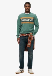 Zielony sweatshirt z logo "Superdry", koszula w kratę związana w talii, niebieskie dżinsy i białe sneakersy z czarnymi paskami. Swobodny design stroju.