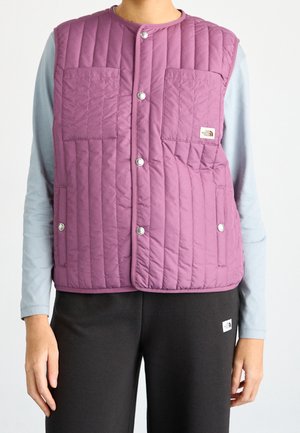 Persoon die een doorgestikt paars mouwloos vest draagt over een lichtblauw shirt met lange mouwen en een zwarte broek, staand tegen een effen achtergrond.